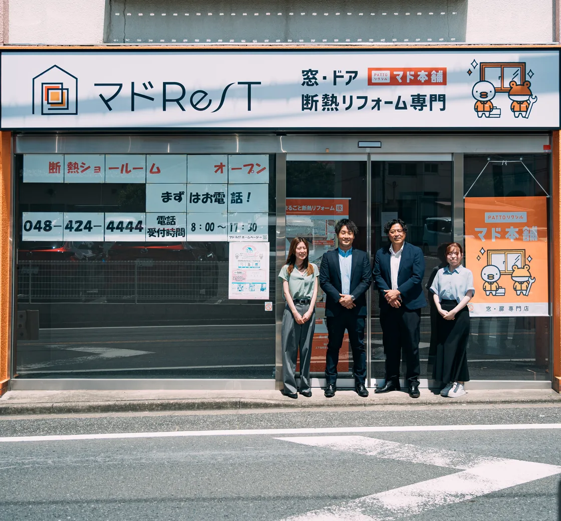 「窓・ドア 断熱リフォーム専門」の看板を掲げたマドレスト朝霞ショールームの外観と、店前に並ぶスタッフ4名